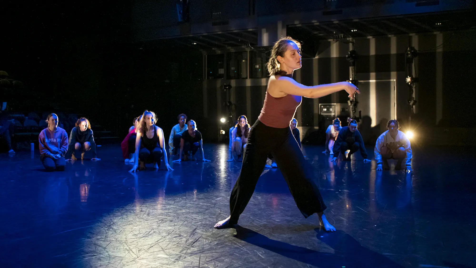 A barefoot dancer performs in the foreground of a dark stage, while a group of dancers crouch in the background under blue lighting.