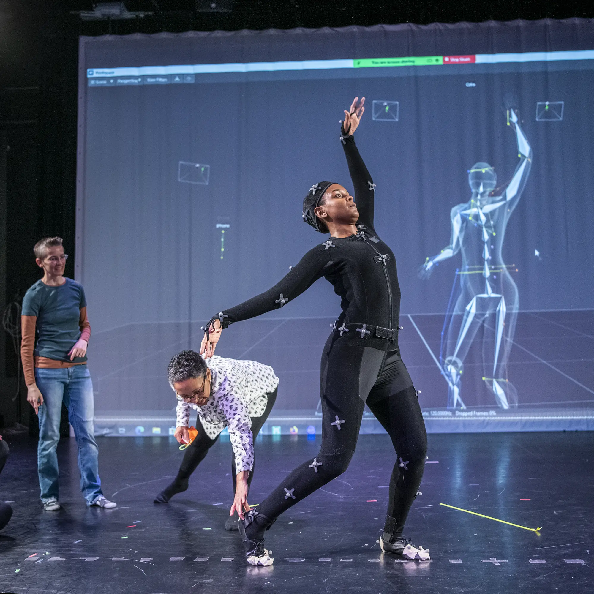A performer in a black motion capture suit strikes a pose in a studio, mirrored exactly by a digital skeletal avatar on a large projection screen behind them, while two people observe and assist.