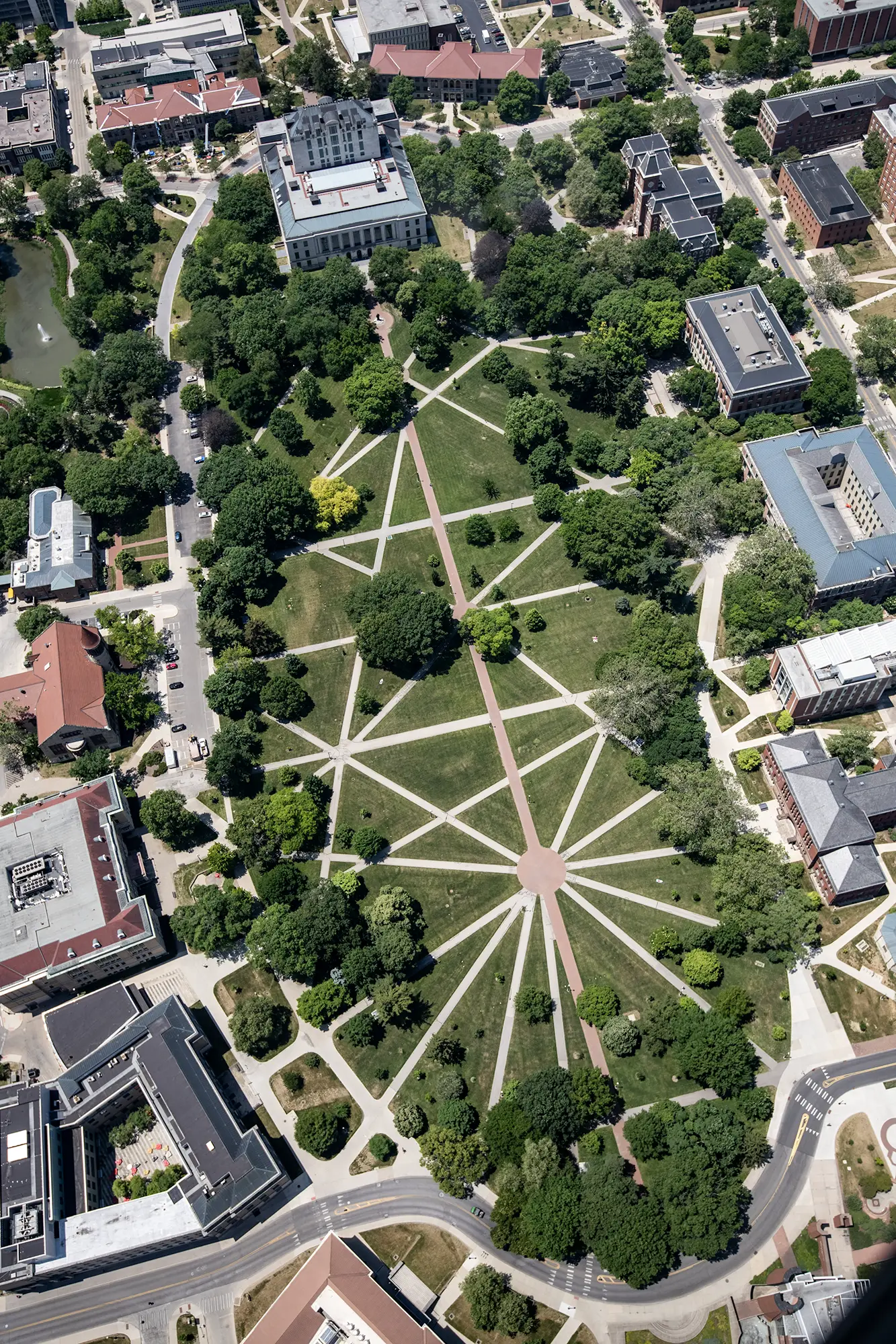 Aerial view of a large, grassy university campus quadrangle crisscrossed by a complex network of intersecting geometric pathways, surrounded by trees and academic buildings.