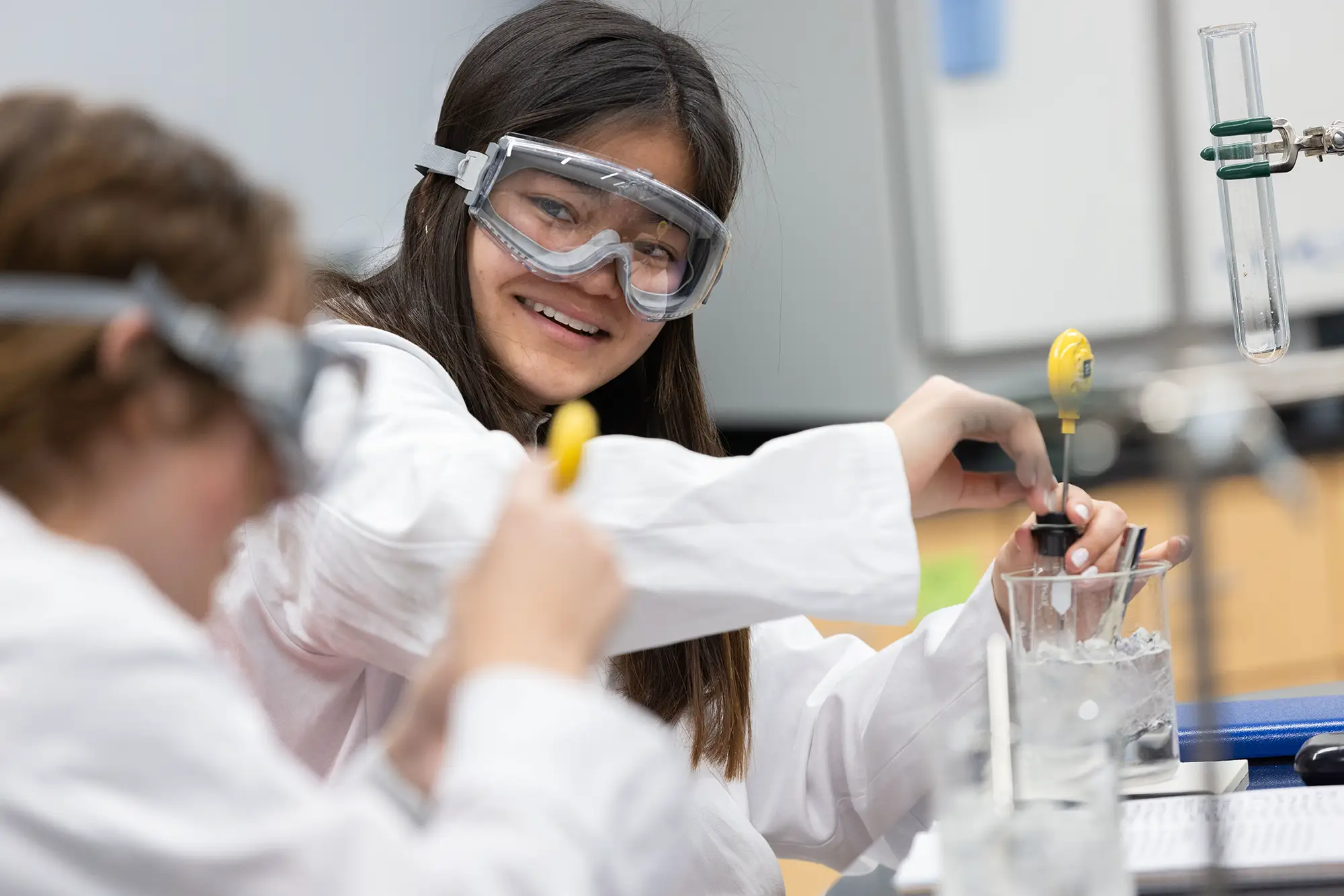 A smiling student wearing safety goggles and a lab coat adjusts a thermometer in an ice bath during a science experiment with a classmate.