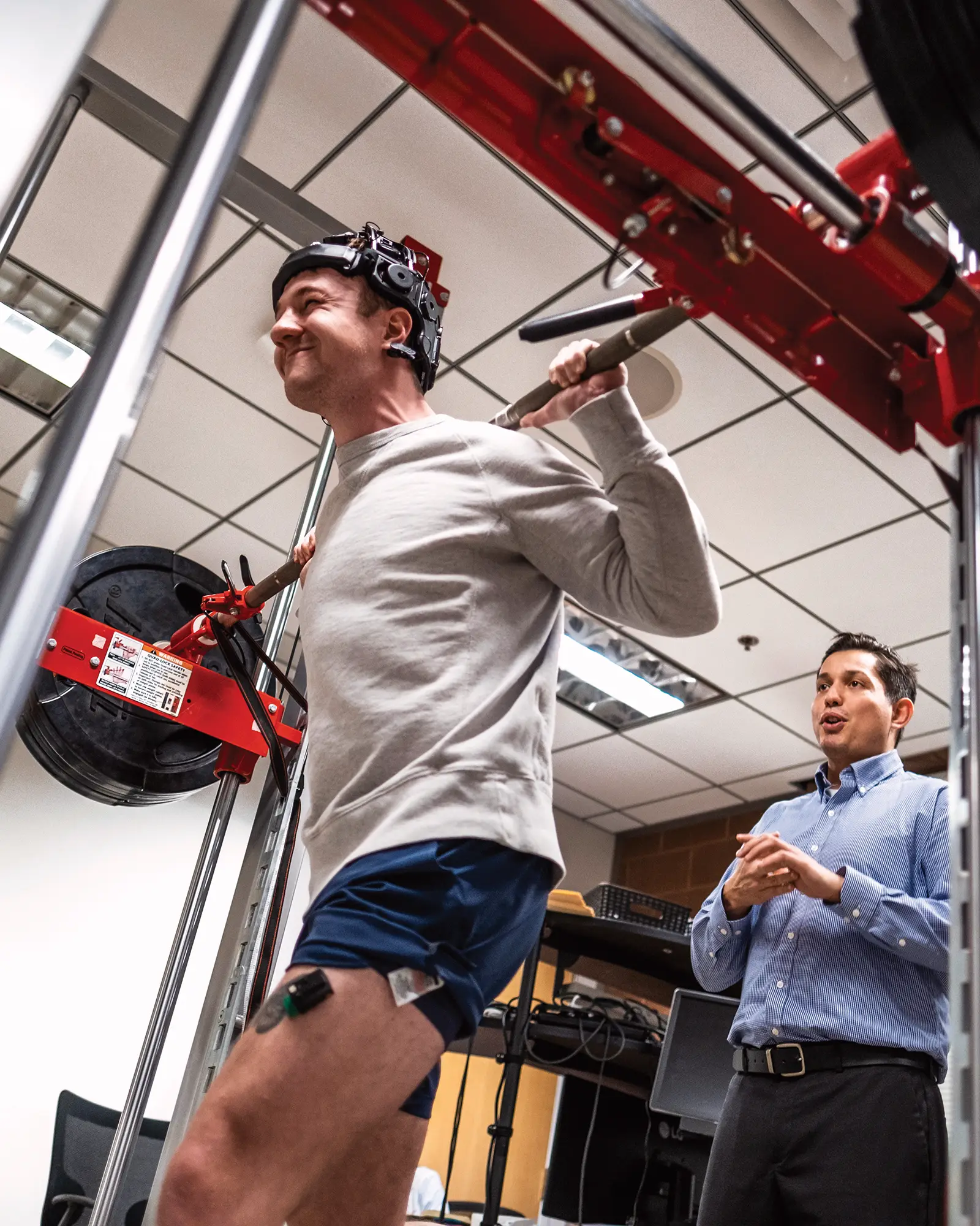 A man wearing a head-mounted sensor cap and a thigh sensor performs a barbell squat on a machine while another man observes him in a lab setting.